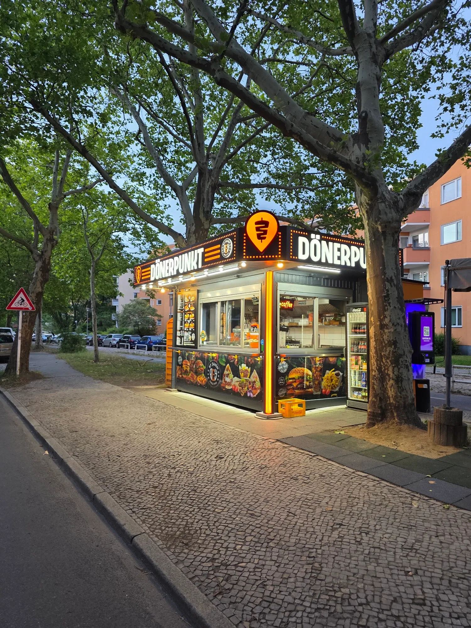 Abendliche Außenansicht des Dönerpunkt Kiosks an der Gallwitzallee unter Bäumen